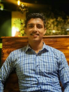 A friendly man in a checkered shirt smiling at a cozy cafe setting.